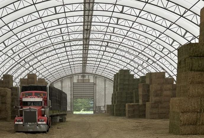 An 18-wheeler semi-truck loading or unloading hay bales inside a high-clearance fabric storage building.